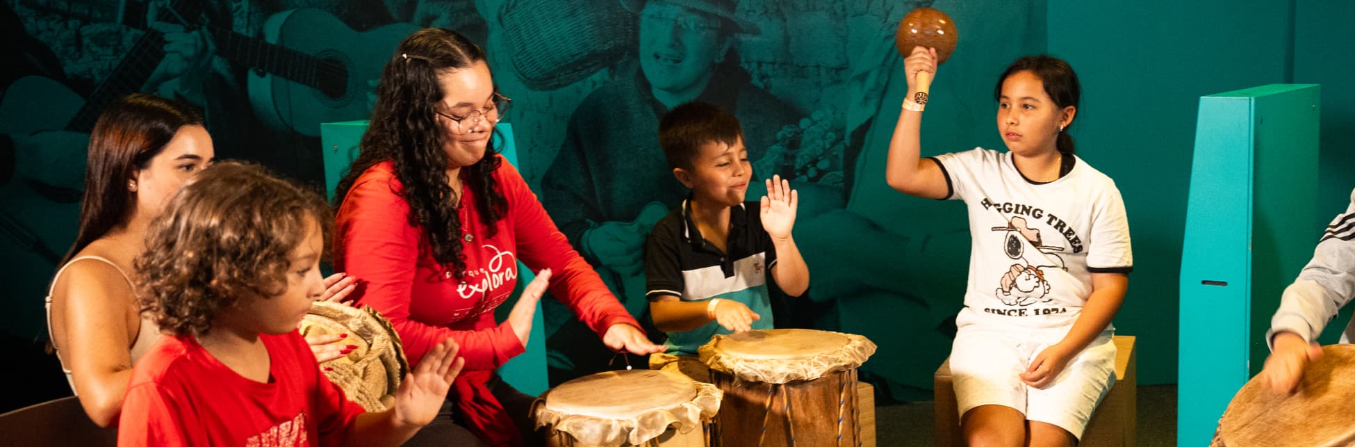Fotografía. Un grupo de niños y jóvenes participa en una actividad musical en la Sala Música del Parque Explora, tocando tambores e instrumentos de percusión en un espacio cerrado con paredes verdes. Una niña sostiene una maraca en alto, mientras los demás siguen el ritmo con las manos. Al fondo, una imagen impresa muestra músicos tocando guitarras.