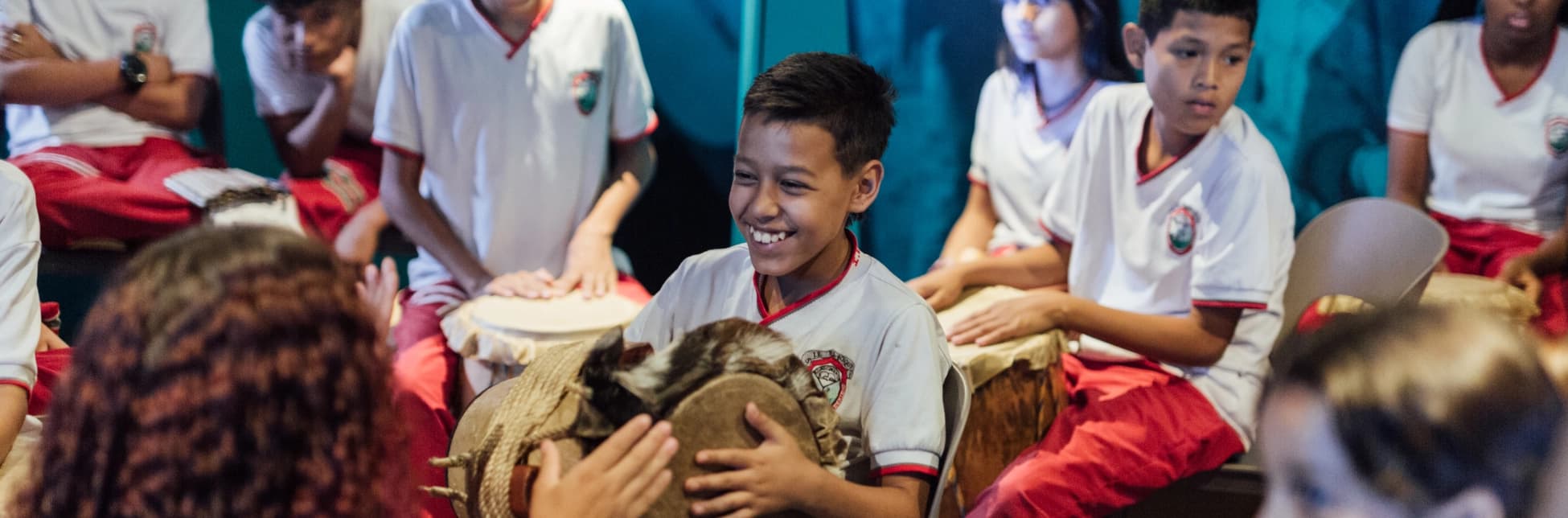 Fotografía. En un espacio interior, varias niñas y niños con uniforme escolar participan en una actividad musical. En el centro, un niño sonríe mientras sostiene un tambor entre sus manos. A su alrededor, otros estudiantes están sentados y tocan tambores de distintos tamaños. En primer plano se observan las cabezas desenfocadas de dos participantes, mientras al fondo hay más estudiantes y una pared decorada con telas.