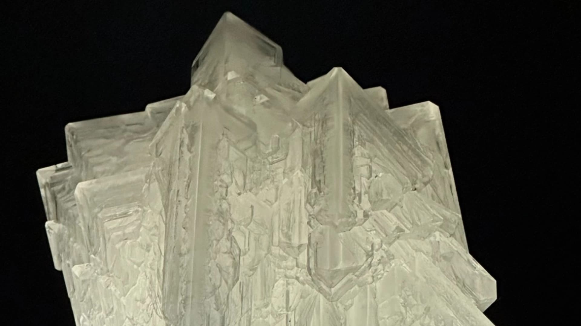 Fotografía. Primer plano de un conjunto de cristales translúcidos sobre un fondo oscuro. Las formaciones tienen superficies lisas y angulares, con bordes rectos y puntas pronunciadas. Varios cristales se superponen y forman una estructura compacta e irregular, con reflejos y zonas de distinta transparencia que permiten apreciar líneas, fracturas y otras formas en su interior.